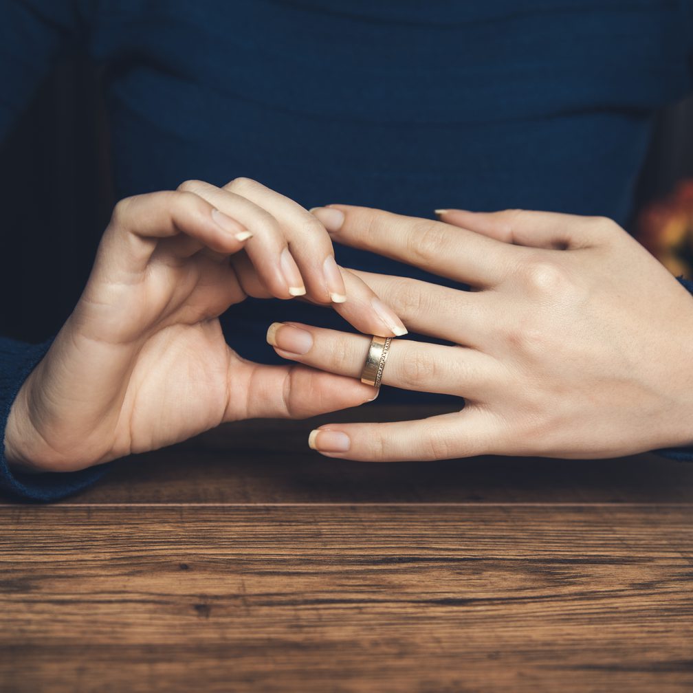 Hands resting on a wooden surface, with one hand touching a wedding ring on the other finger, wearing a dark blue long-sleeve shirt.