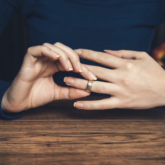 Hands resting on a wooden surface, with one hand touching a wedding ring on the other finger, wearing a dark blue long-sleeve shirt.