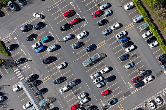 Aerial view of a parking lot filled with numerous cars arranged in diagonal rows, with marked parking bays, pedestrian crossings and surrounding greenery.