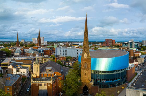 Panoramic cityscape featuring Gothic spire church in foreground, blue cylindrical building, and a mix of brick and glass structures under a cloudy sky.