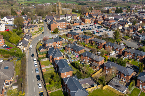 Aerial view of a UK town with red-brick houses, a tall church tower, winding streets, a central parking area, and green fields dotted with trees.