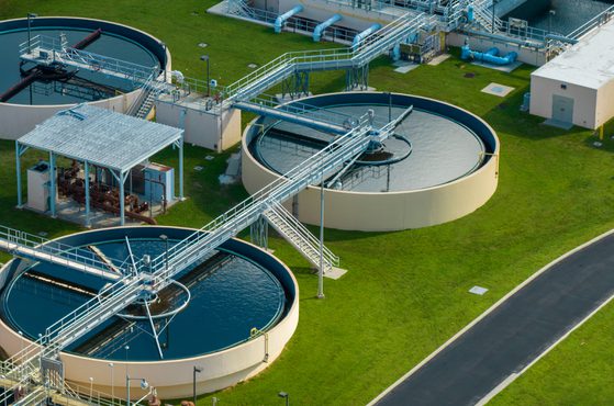 Aerial view of a wastewater treatment plant with large circular tanks, metal walkways, and pipes over green grass and paved pathways.