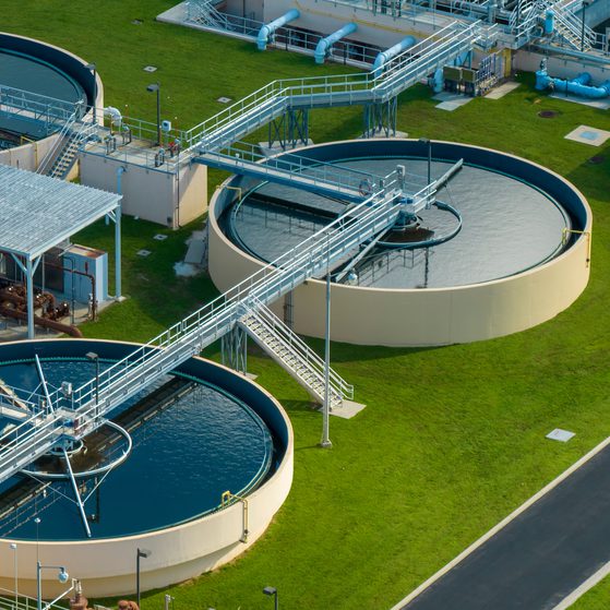 Aerial view of a wastewater treatment plant with large circular tanks, metal walkways, and pipes over green grass and paved pathways.