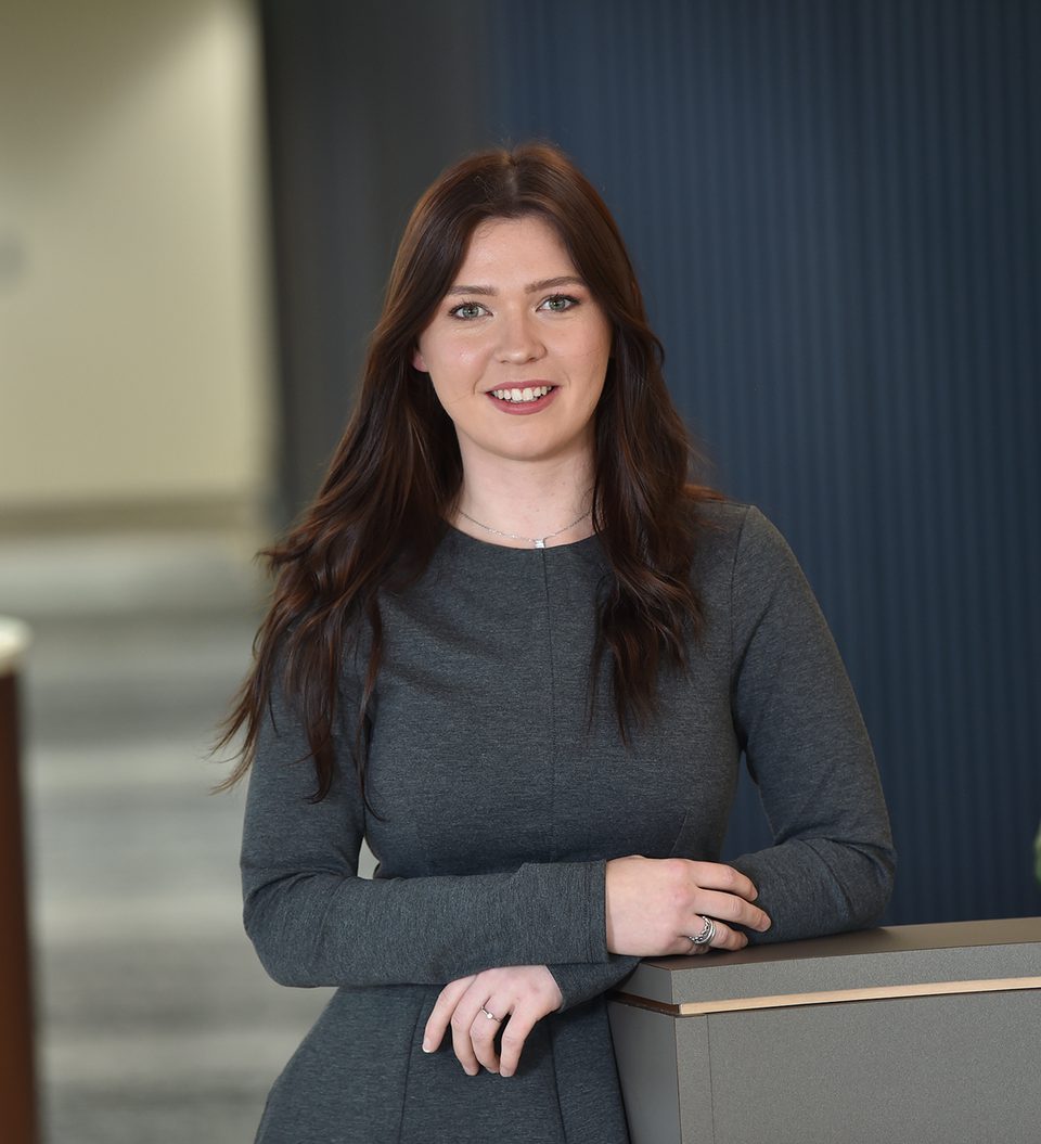 A young woman with long brown hair, wearing a grey long-sleeve dress, stands casually at a reception desk with a friendly smile in a modern office environment.