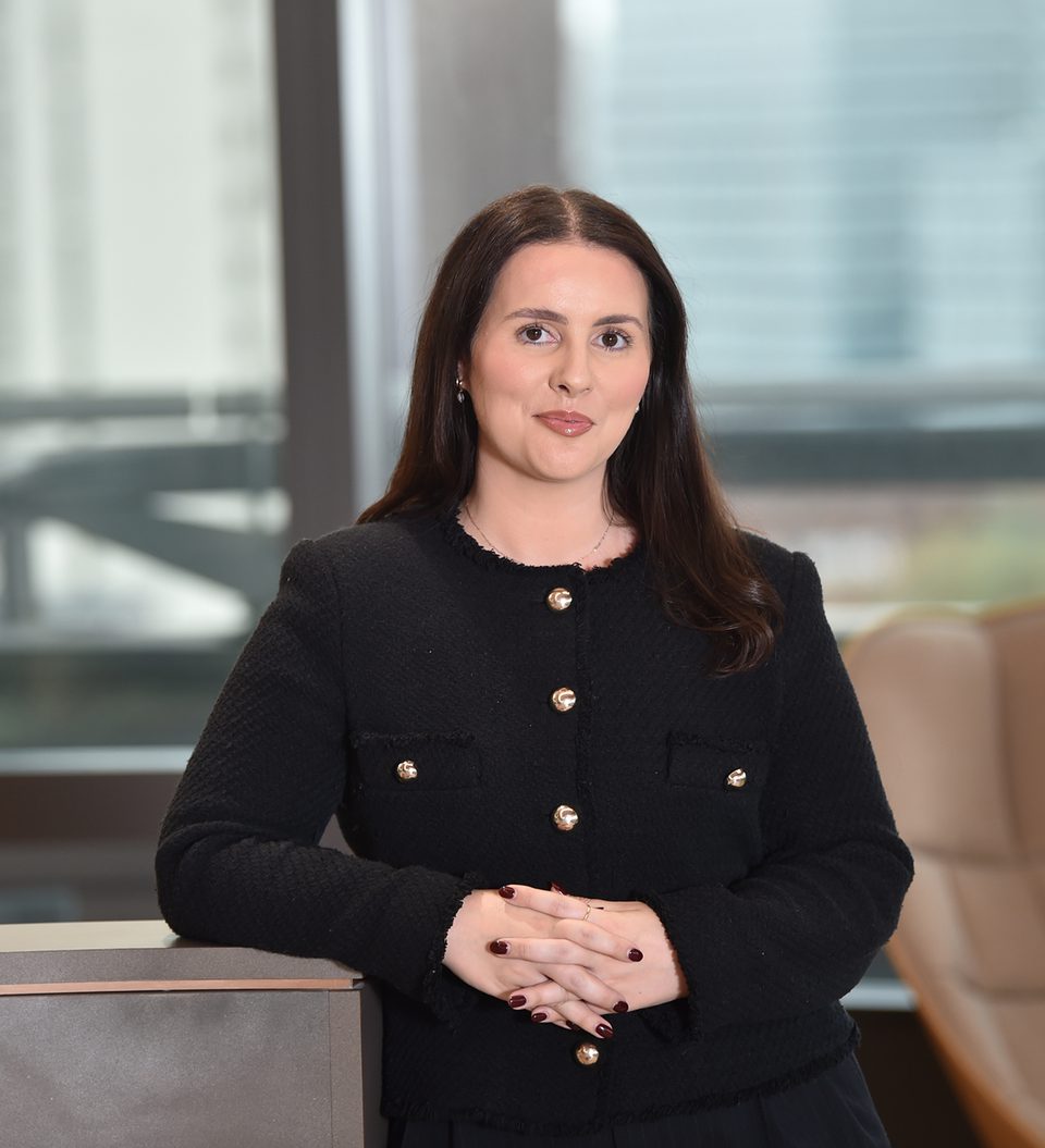 A woman with dark brown hair, dressed in a black blazer with gold buttons, standing with hands clasped in front of a desk in a modern office.
