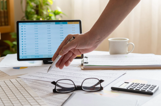 A hand uses a pen over documents on a bright desk, beside a laptop with a spreadsheet, glasses, calculator, clipboard, mug and a plant nearby.