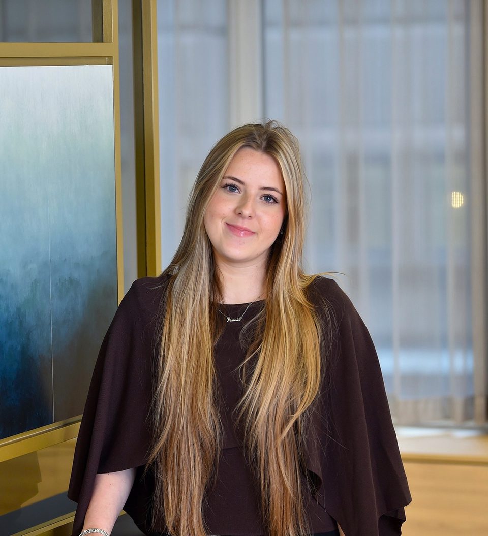 A young woman with long blonde hair, smiling and wearing a black graduation gown, standing indoors next to large windows.