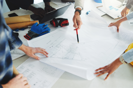 Several people collaborate over architectural blueprints on a table with rolled plans, a yellow hard hat, gloves, tools and a laptop as a pencil points at the drawings.