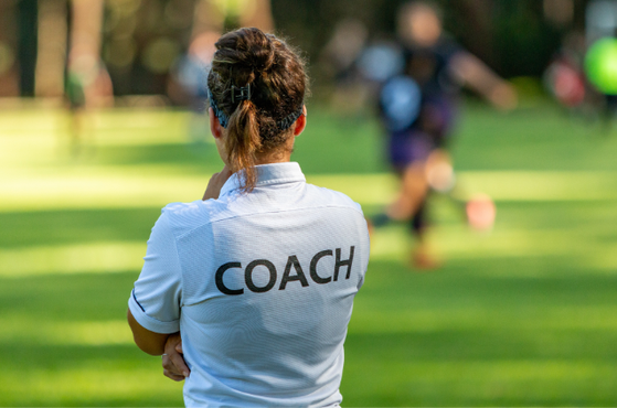 Back view of a person in a white polo with COACH on the back, standing on a sunlit grassy field while blurred players train in the background.