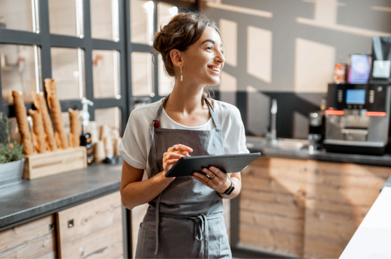 A person in a grey apron stands behind a bakery counter, smiling as they hold a tablet, with baguettes and a coffee machine visible in the background.