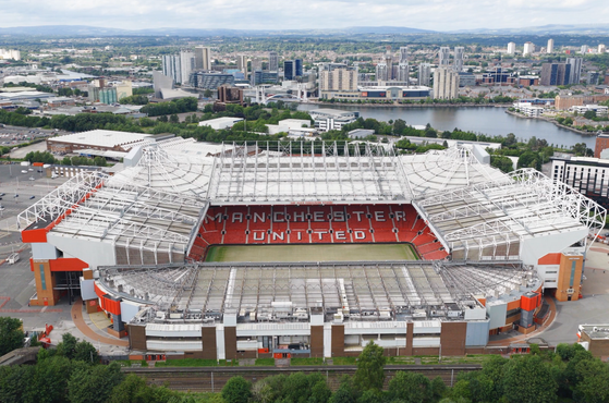 Aerial view of Old Trafford stadium with red seats spelling MANCHESTER UNITED, white lattice roof, and surrounding cityscape with a river and distant hills.