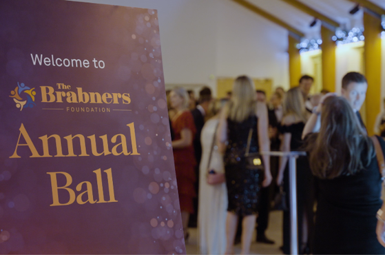 Purple banner in the foreground reads 'Annual Ball' with The Brabners Foundation logo; a crowd of guests mingles in a brightly lit event hall in the background.