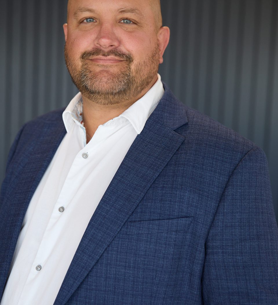 A bald person with blue eyes and a light beard, wearing a white shirt and navy blazer, posing in front of a dark vertical-panel backdrop.