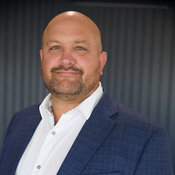 bald person with blue eyes and a short beard, wearing a white shirt and navy blazer, smiling subtly against a dark vertical-panel background