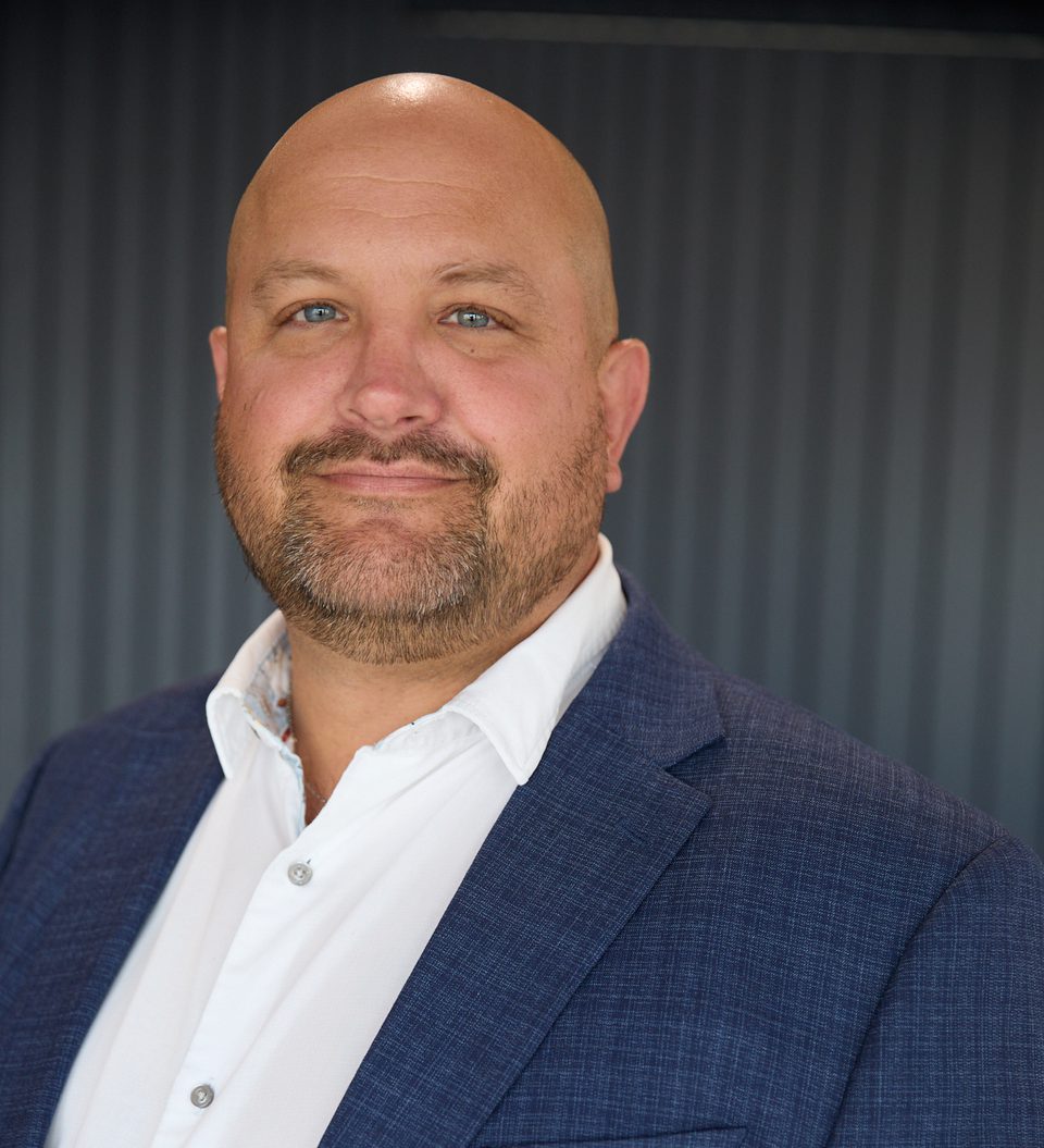 bald person with blue eyes and a short beard, wearing a white shirt and navy blazer, smiling subtly against a dark vertical-panel background