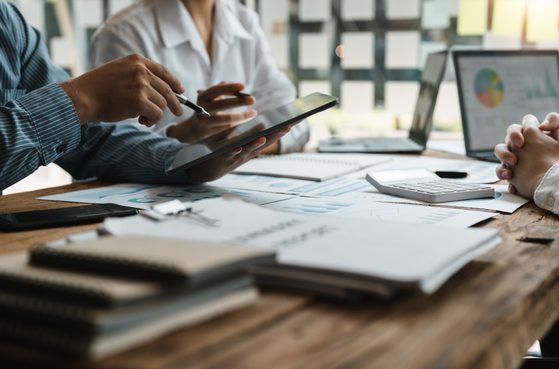 People in a meeting at a wooden table with documents, laptops, and a tablet, engaged in discussion, with sunlight streaming in through large windows.