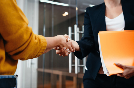Two people shake hands in a modern office setting, one holding an orange folder while the other wears a mustard yellow sweater.