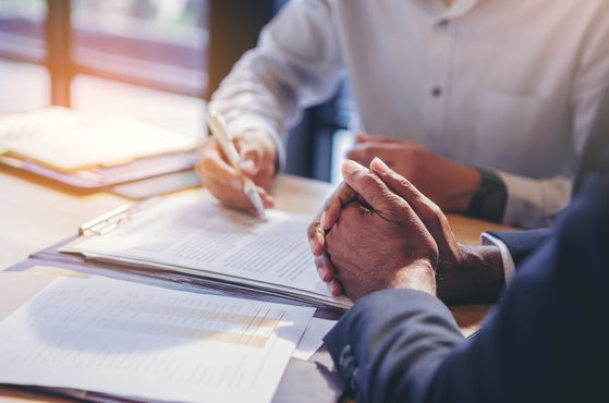 Two people in business attire sitting at a table, one with clasped hands and the other writing on a document, in a bright office setting.