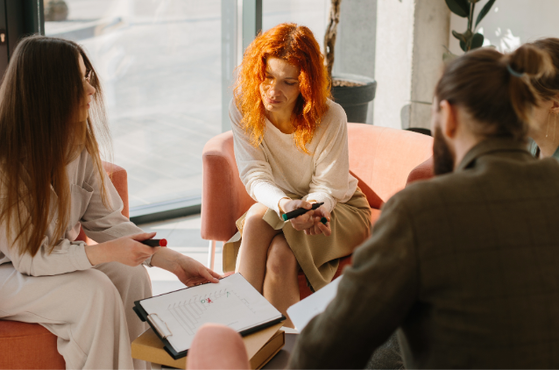 Four people sit on pink chairs in a bright room with large windows; they are engaged in a discussion, with notebooks and phones on their laps.