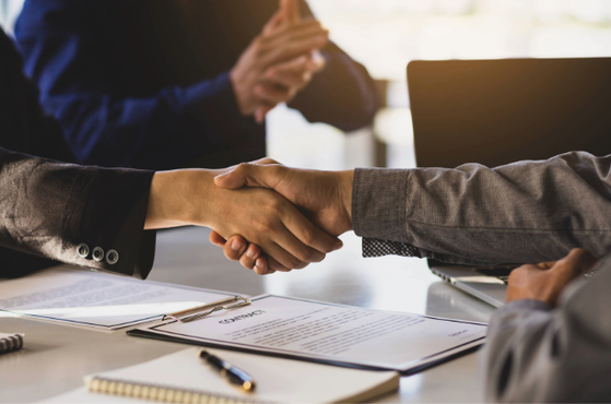 Two people in business attire shake hands across a table, with documents, a pen, and a laptop visible in a professional meeting setting.