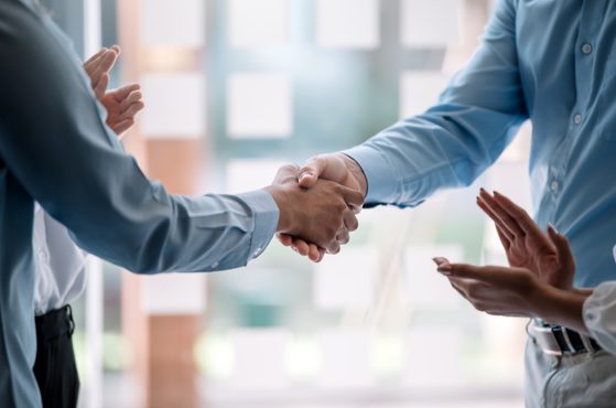 Two people are shaking hands in a professional setting, with a third person clapping nearby. They are wearing formal shirts, with a blurred office background.
