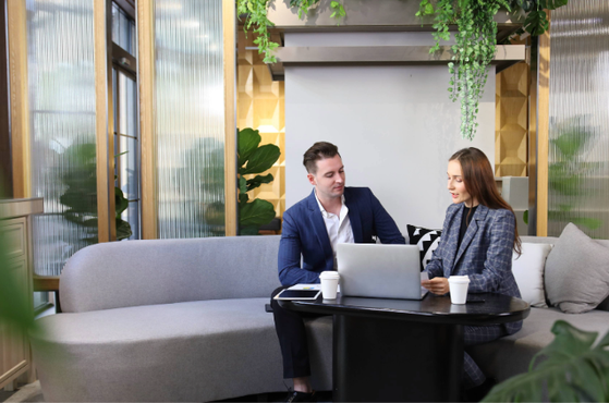 A man and woman sitting on a grey sofa in a modern office lounge, looking at a tablet together with a laptop and two coffee cups on the black table.