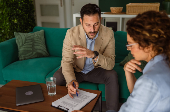 A man and woman sit on a green sofa engaged in a serious discussion, with documents and a laptop on a wooden coffee table in front of them.