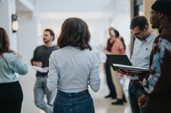 A group of people engaged in conversation in a modern indoor space with natural light, some holding notebooks or laptops, and blurred background figures.