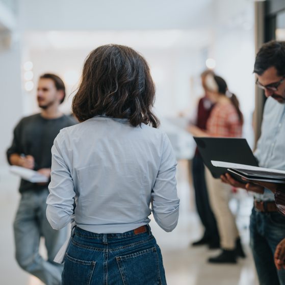 A group of people engaged in conversation in a modern indoor space with natural light, some holding notebooks or laptops, and blurred background figures.
