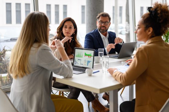 Four professionals sit around a white table in a modern office, engaged in a meeting. Two laptops display charts and graphs, and glasses of water are on the table.