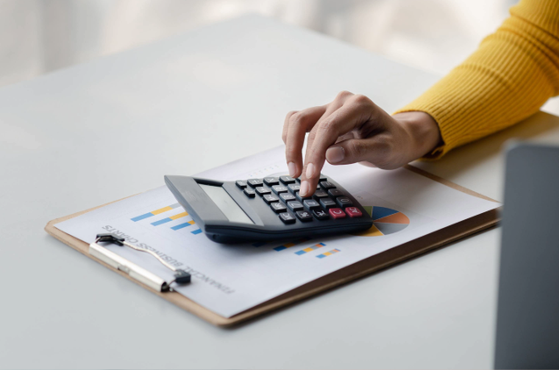 A person in a yellow jumper using a calculator on a desk with financial charts and a clipboard, with part of a laptop visible.