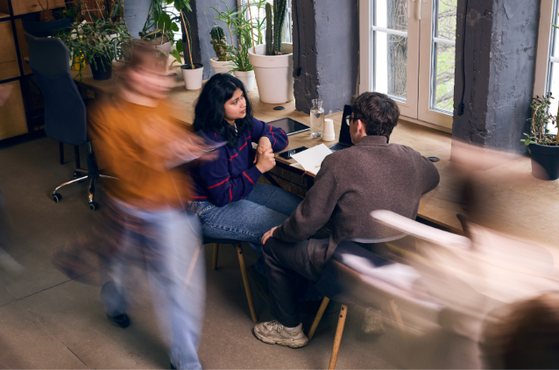 Two people sit at a wooden table by a large window, talking, while blurred passers-by move through a plant-filled, sunlit workspace with coffee mugs on the table.