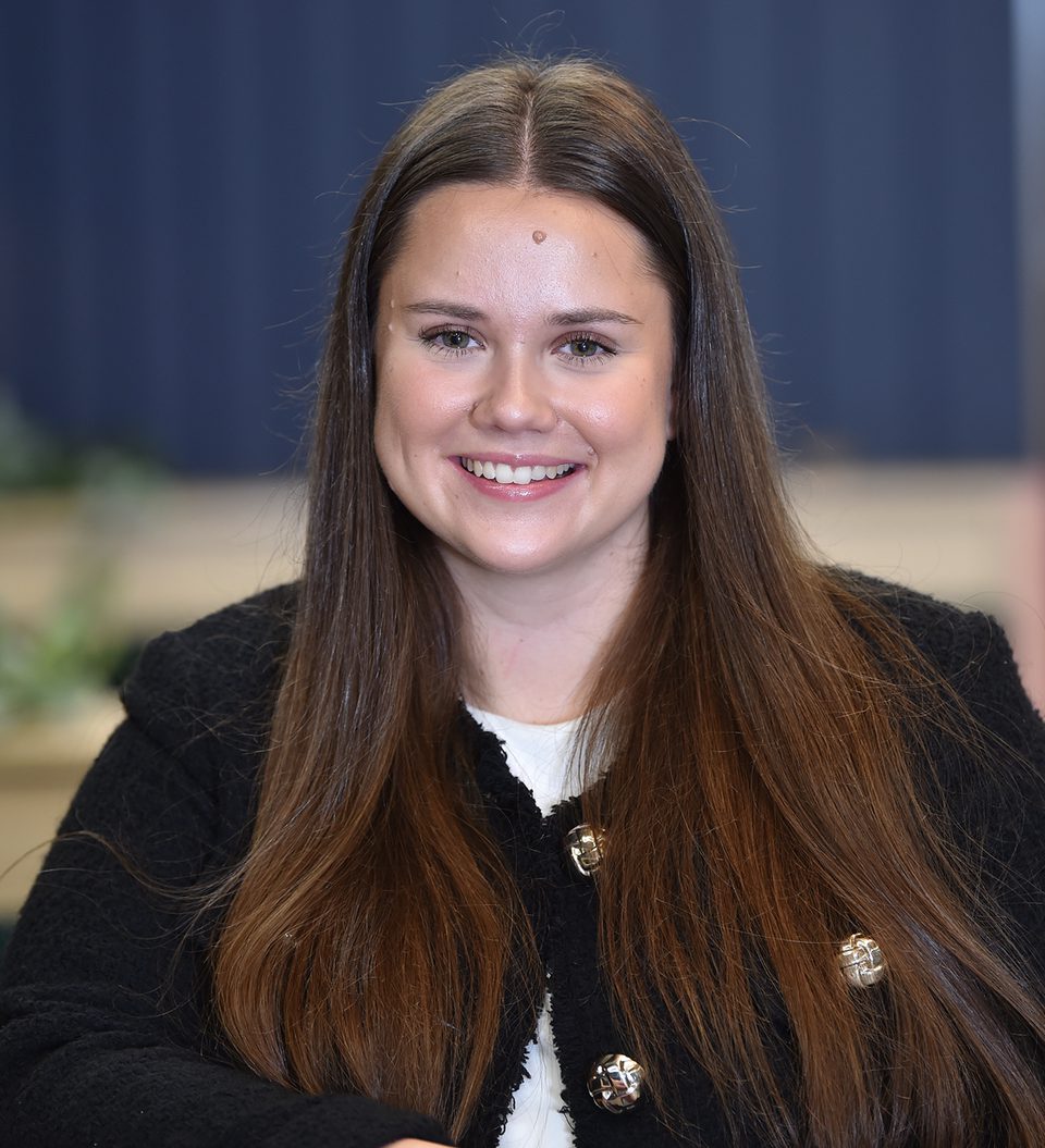 A young woman with long brown hair, smiling and wearing a black cardigan with gold buttons, sitting indoors with a blurred background.
