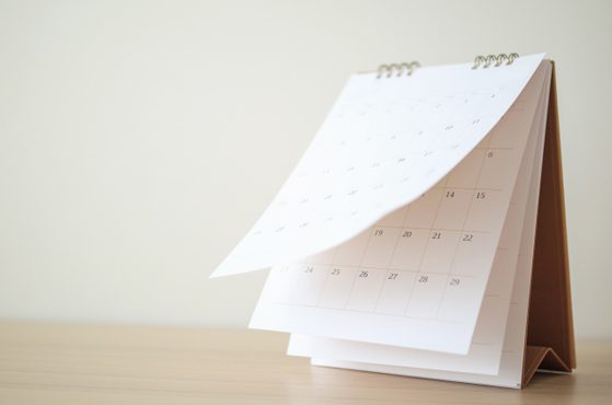 Desk calendar on a light wooden desk with a spiral binding, pages fanning out and displaying dates, supported by a brown stand against a pale wall.