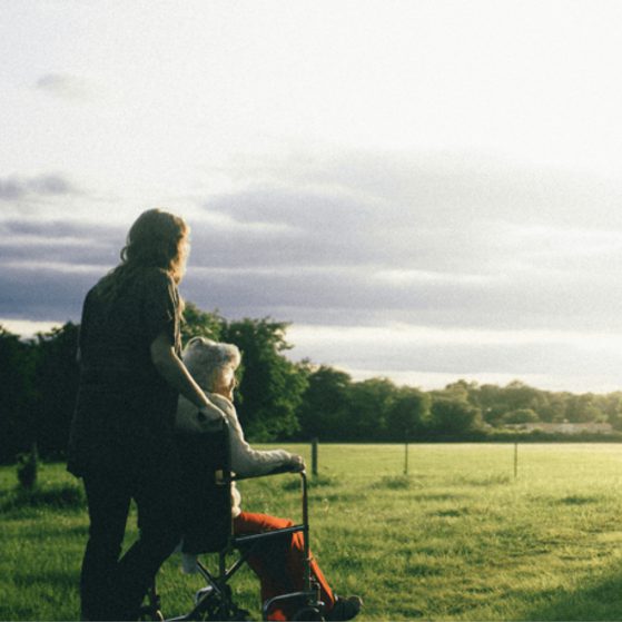 A woman with a young child in a wheelchair walk together in a sunny, open grassy field with trees and a fence in the distance.