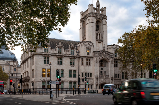 Supreme Court on a busy London street, framed by autumn trees; a red double-decker bus and cars pass by.