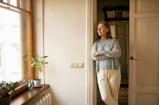 A woman stands with arms crossed, gazing out of a window in a softly lit room with cream walls and wooden accents.