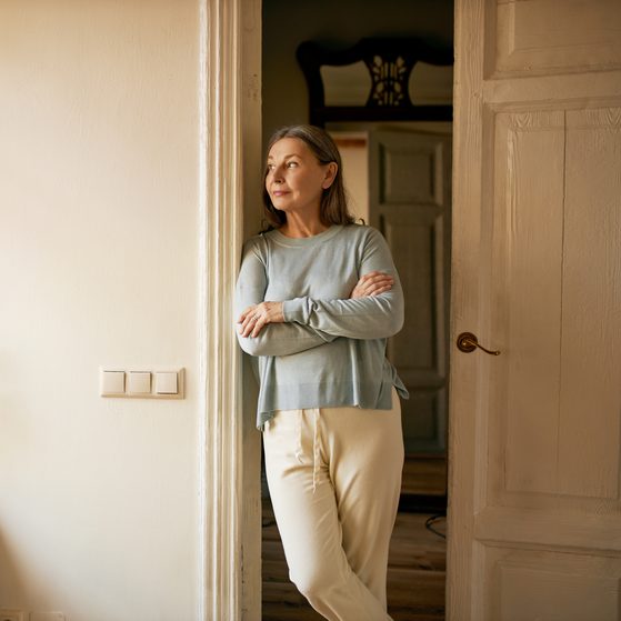 A woman stands with arms crossed, gazing out of a window in a softly lit room with cream walls and wooden accents.