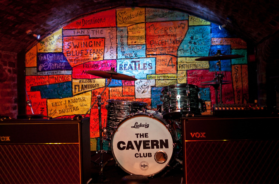 Dim cave-like stage with a Ludwig drum kit reading The Cavern Club, flanked by two VOX amplifiers, and a vibrant wall of colorful band-name patches.