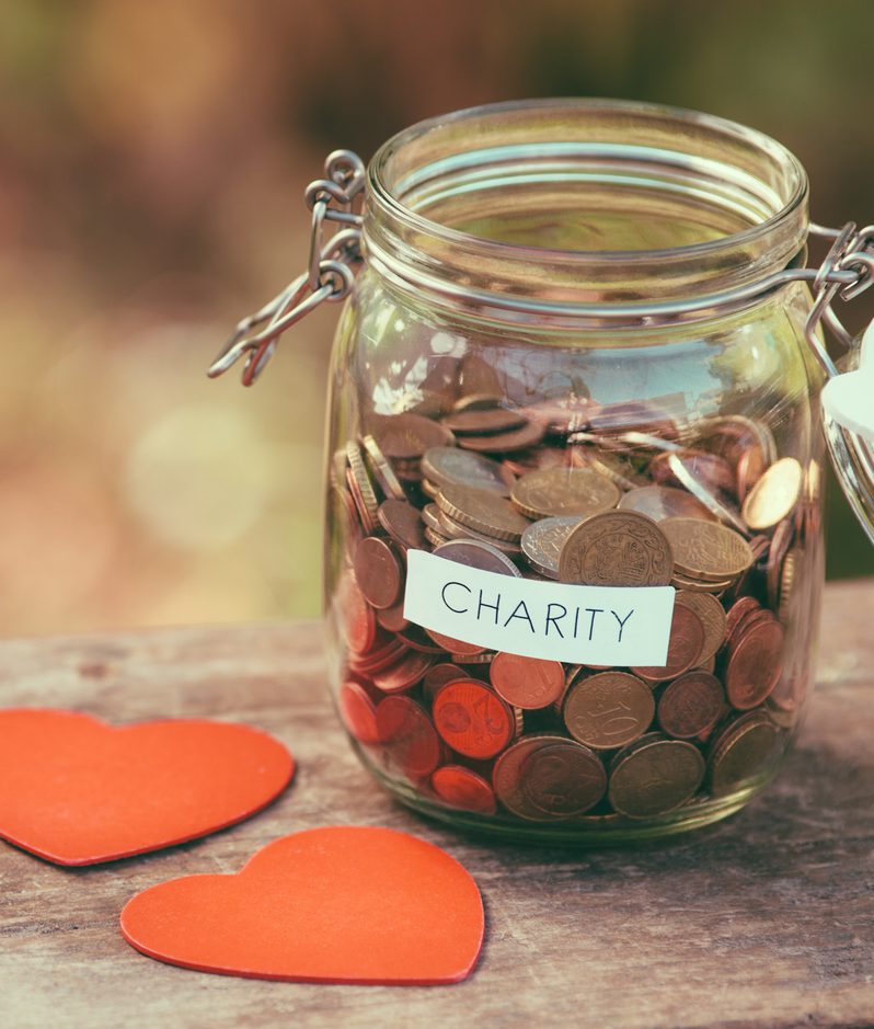 A glass jar filled with assorted coins, labelled "CHARITY," is placed on a wooden surface next to two red paper hearts. The background is blurred with warm tones.