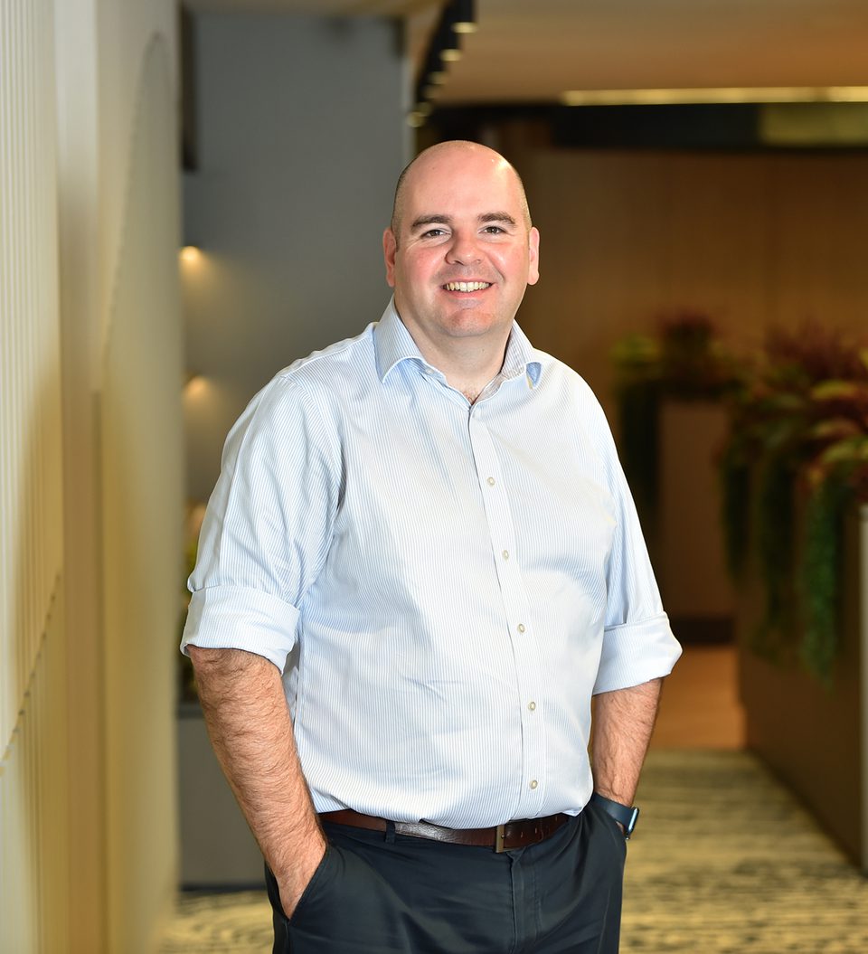 A smiling man with a shaved head wearing a light blue shirt and dark trousers, standing indoors with hands in pockets against a background of plants and warm lighting.