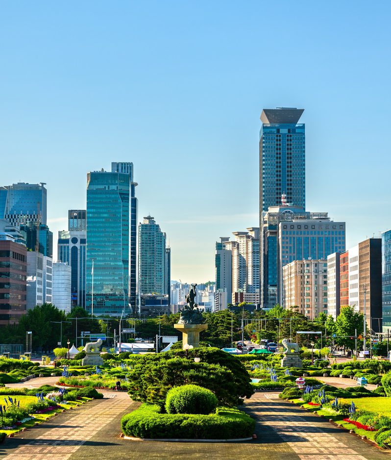 Urban skyline of glass towers behind a landscaped park with a central statue fountain, trimmed hedges and vibrant flower beds under a bright blue sky.