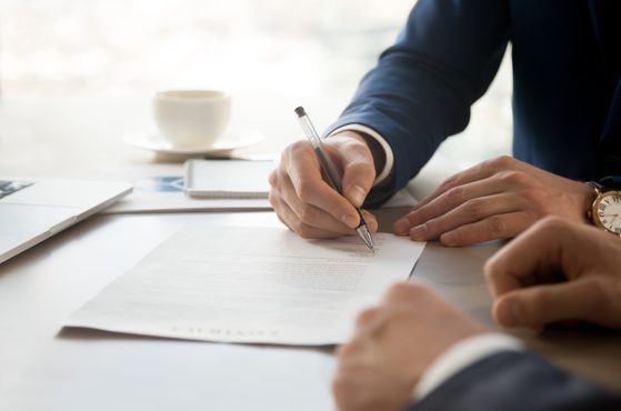 A person in a dark suit signs a document on a white table, with a cup and notebook in the background, during a professional or business meeting.