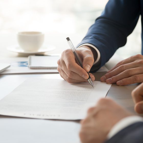 A person in a dark suit signs a document on a white table, with a cup and notebook in the background, during a professional or business meeting.