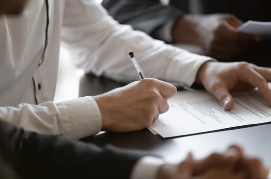 Person signing a document with a pen on a table, with another individual next to them reading a paper. Focus on hands and documents, background is blurred.