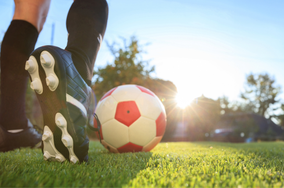 Close-up of a black football boot with studs beside a red‑and‑white soccer ball on sunlit green grass, with a bright sky and distant trees in the background.