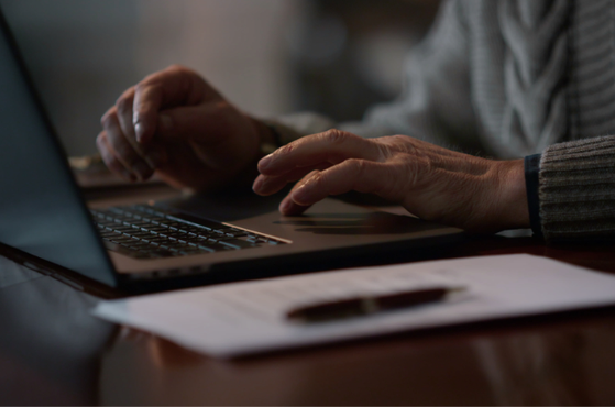 Close-up of hands typing on a laptop at a wooden desk, with a sheet of paper and a pen in the foreground; the user wears a grey knitted sweater.