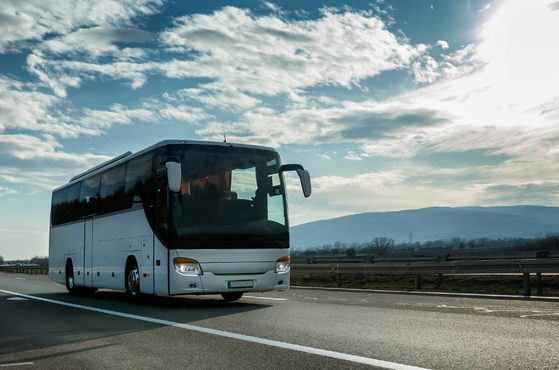 White touring coach on a highway with metal guard rails, under a partly cloudy sky, distant hills on the horizon and sunlight on the right.