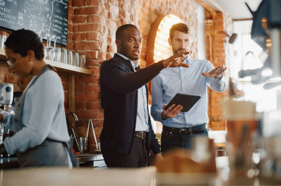 Two men are having a discussion behind the counter of a coffee shop, with one holding a tablet and gesturing. A woman is preparing drinks nearby.
