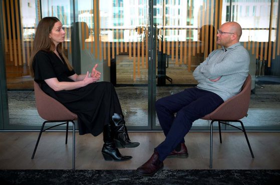 A woman in a black dress and tall black boots sits facing a bald man in a grey sweater and dark pants; they are in a modern office space with glass walls behind them.